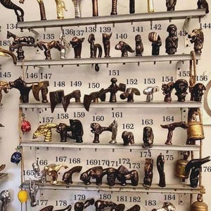 K&ouml;nnte beinhalten: Eine Sammlung dekorativer T&uuml;rgriffe und -kn&ouml;pfe, ausgestellt auf Metallregalen. Die Griffe weisen verschiedene Designs auf, darunter Tierfiguren und geometrische Formen. Die Farbpalette umfasst Bronze-, Gold- und Dunkelbraunt&ouml;ne. Ma&szlig;e in cm.