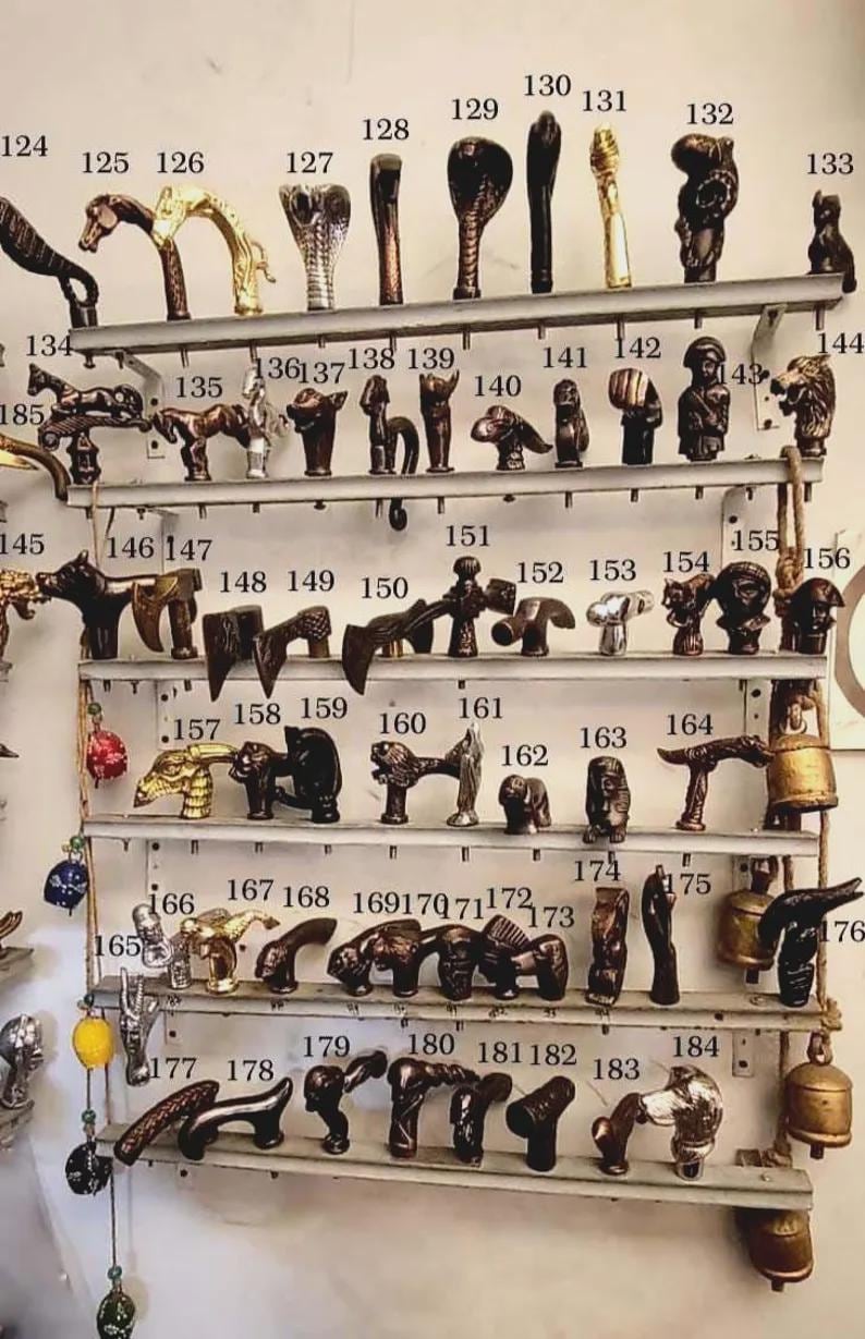 K&ouml;nnte beinhalten: Eine Sammlung dekorativer T&uuml;rgriffe und -kn&ouml;pfe, ausgestellt auf Metallregalen. Die Griffe weisen verschiedene Designs auf, darunter Tierfiguren und geometrische Formen. Die Farbpalette umfasst Bronze-, Gold- und Dunkelbraunt&ouml;ne. Ma&szlig;e in cm.