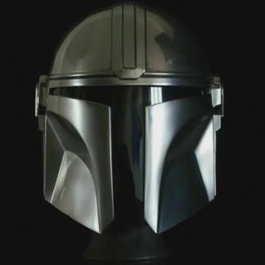 May include: A metallic silver helmet with a distinctive T-shaped visor and angular features. The helmet has a smooth, reflective surface and a dark background, showcasing its sleek design. This helmet is a replica of the Mandalorian helmet.