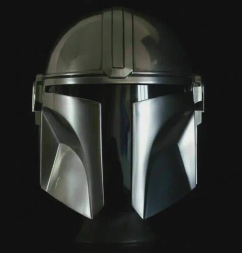 May include: A metallic silver helmet with a distinctive T-shaped visor and angular features. The helmet has a smooth, reflective surface and a dark background, showcasing its sleek design. This helmet is a replica of the Mandalorian helmet.