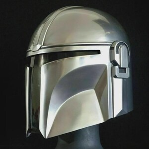 May include: A polished silver Mandalorian helmet, featuring a T-shaped visor and a distinctive, angular design. The helmet has a smooth, reflective surface and is set against a dark background. The helmet is a replica of the one worn by the Mandalorian.