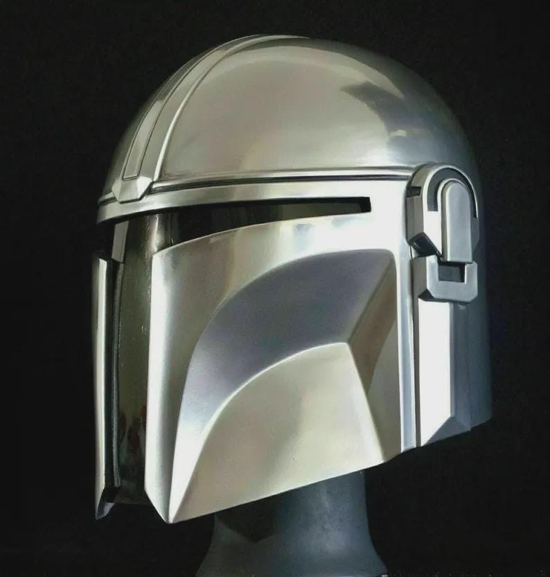 May include: A polished silver Mandalorian helmet, featuring a T-shaped visor and a distinctive, angular design. The helmet has a smooth, reflective surface and is set against a dark background. The helmet is a replica of the one worn by the Mandalorian.