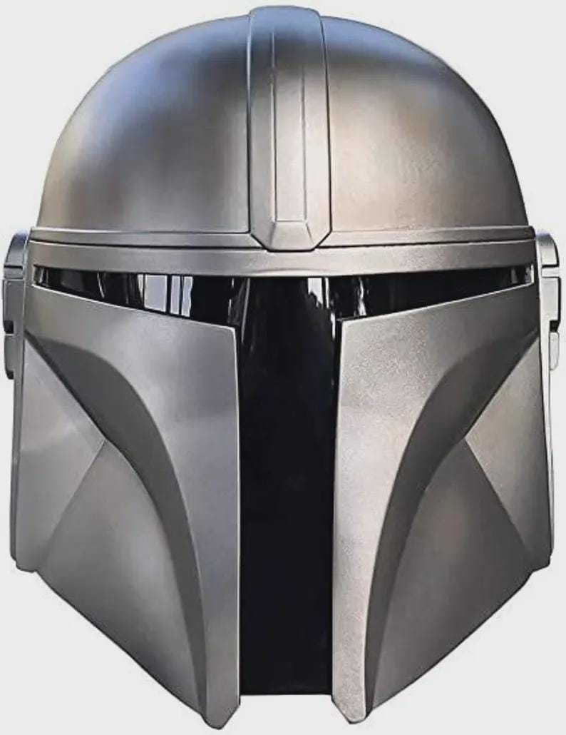 May include: A metallic silver Mandalorian helmet with a distinctive T-shaped visor and a rounded top. The helmet features angular cheek plates and a central vertical ridge, showcasing a sleek, futuristic design. The helmet is a replica of the one worn by the Mandalorian.