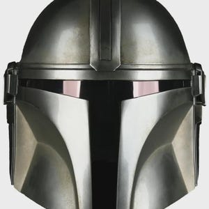 May include: A close-up of a silver Mandalorian helmet. The helmet has a rounded top with a vertical ridge and a visor with a dark, reflective surface. The lower face area is angular and metallic.
