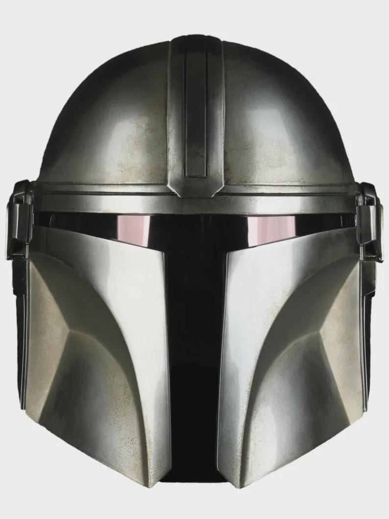 May include: A close-up of a silver Mandalorian helmet. The helmet has a rounded top with a vertical ridge and a visor with a dark, reflective surface. The lower face area is angular and metallic.