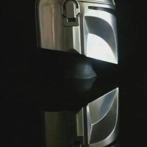 May include: A shiny, silver Mandalorian helmet, reflecting light against a dark background. The helmet features a T-shaped visor and a distinctive, angular design, typical of the Star Wars character.