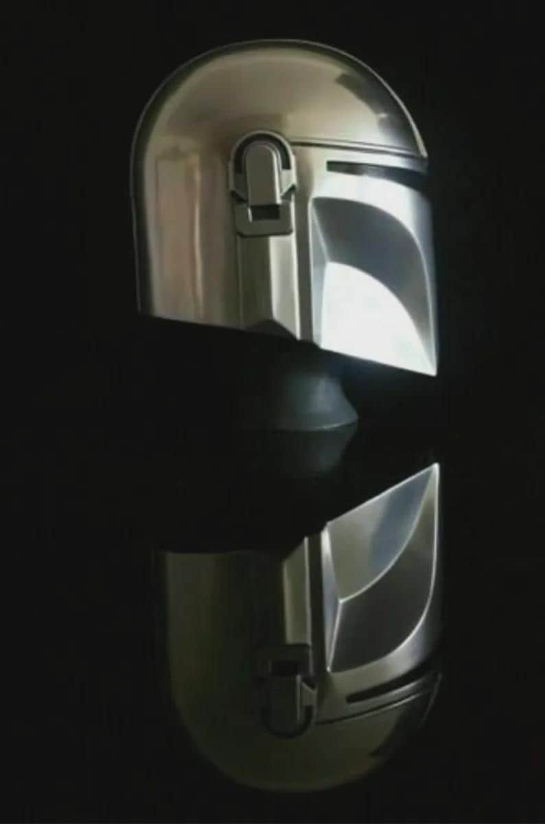 May include: A shiny, silver Mandalorian helmet, reflecting light against a dark background. The helmet features a T-shaped visor and a distinctive, angular design, typical of the Star Wars character.