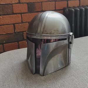 May include: A silver Mandalorian helmet with a visor, resting on a gray surface. The helmet has a rounded top and angular features, with visible wear and tear. The background includes a brick wall and a radiator.