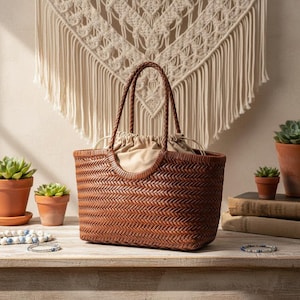 Könnte beinhalten: Eine braune, gewebte Ledertasche mit geflochtenen Henkeln. Die Tasche hat ein beiges Futter mit Kordelzug. Kleine Topfpflanzen und Armbänder sind ebenfalls zu sehen.