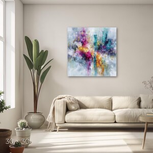 May include: A square abstract painting, 30 x 30 inches, with a vibrant display of colors. The artwork features a blend of pink, yellow, teal, and purple hues, creating a dynamic and expressive composition. The painting is mounted on a white wall above a beige sofa.