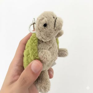 May include: A plush turtle keychain with a light green shell and tan body. The turtle has a small black eye and a silver metal keyring. The soft toy is held against a white background.