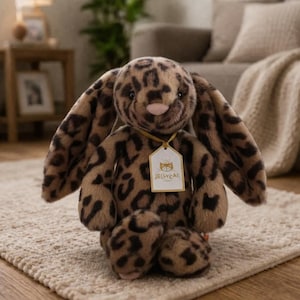 May include: A plush bunny toy with a leopard print pattern in shades of brown and tan. The bunny has long floppy ears, a pink nose, and a tag that reads "Jellycat". The toy is sitting on a beige rug.
