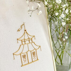 May include: A framed artwork features a gold-colored pagoda design embroidered on a cream-colored canvas. The pagoda has multiple tiers and a flag at the top. The frame is gold, and a vase of white flowers is in the background.