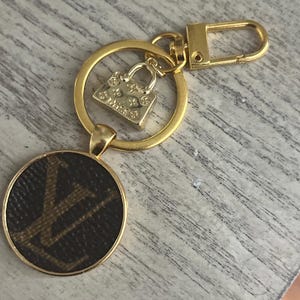 May include: Gold-toned keychain with a round pendant featuring a brown and tan pattern. A small gold purse charm and a gold ring with a clasp are also attached. The keychain is designed for keys or accessories.