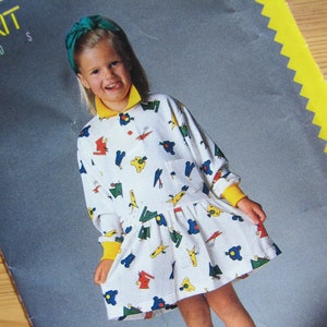 May include: A young girl wearing a white dress with a colorful geometric pattern. The dress has a yellow collar and a green bow in her hair. She is wearing black shoes and white socks.