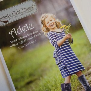 May include: A sewing pattern envelope for the Adele knit dress by Violette Field Threads. The cover features a child wearing a blue and white striped dress with a collared neckline and ruffled back, paired with grey velvet boots, holding yellow flowers.