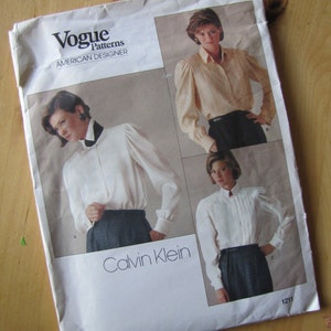 May include: Vogue Patterns American Designer sewing pattern featuring three different designs by Calvin Klein. The pattern is for a white button-down shirt with a collar and long sleeves. The pattern number is 1211.