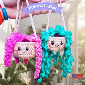 May include: Two crocheted hanging planters with smiling faces, one featuring bright pink trailing vines and the other featuring vibrant teal trailing vines. Each beige pot has black eyes, a small stitched mouth, and pink blush. Text reads PDF PATTERN.