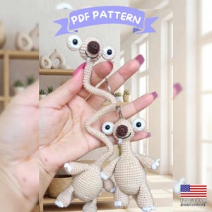 Peut inclure: Un modèle PDF au crochet représentant deux créatures amigurumi beiges ressemblant à des paresseux, avec de longs cous, des nez marron foncé, des dents blanches et de grands yeux ronds. Les figurines sont fixées à des porte-clés.