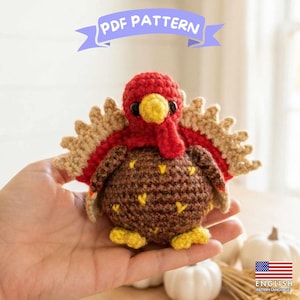 Op de afbeelding: Een gehaakte kalkoen amigurumi met een rond bruin lichaam met gele accenten, een rode kop, een gele snavel en een rode lel. De kalkoen heeft een waaiervormige staart met beige en rode veren. Een blauwe banner bovenaan toont PDF PATTERN.