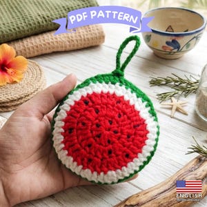 May include: A round, crocheted watermelon-themed scrubby featuring a red center with black seed-like stitches, a white border, and a green outer edge. The item includes a small green loop at the top for hanging and is labeled as a PDF pattern.