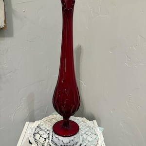 Puede incluir: Un jarrón de cristal rojo rubí alto con un cuello esbelto y alargado que se ensancha en un borde ondulado de múltiples puntas. La base del jarrón tiene un cuerpo bulboso y texturizado con crestas verticales, sobre un pie circular.