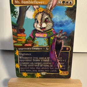 May include: A collectible trading card titled Ms. Bumbleflower featuring a rabbit character with a stitched face, wearing a flower crown and green outfit, holding a tray of colorful donuts. The card text includes game rules and stats 1/5.