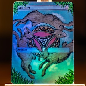 May include: A collectible card featuring a fantasy illustration of three hares leaping around a central design. The card has a blue and green background with the text "Sol Ring" and "Artifact". The artwork is detailed with shades of brown, pink, and green.