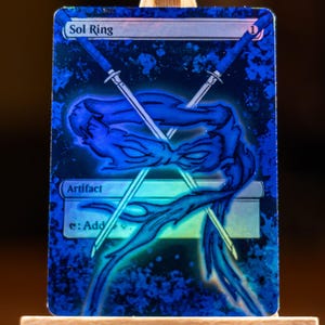 May include: A blue Sol Ring card with a ninja turtle design. The card features two crossed swords and a blue character illustration. The text "Sol Ring" and "Artifact" are visible. The card has a holographic finish.