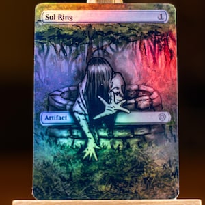 May include: A holographic Sol Ring card featuring a spectral figure emerging from a well. The card has the text "Sol Ring" at the top and "Artifact" at the bottom. The artwork is detailed with a mix of colors.