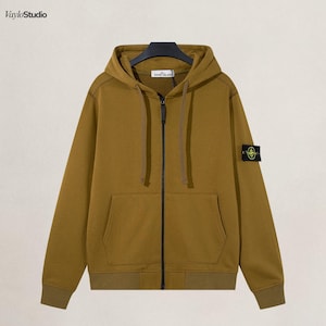 Könnte beinhalten: Senffarbener Kapuzenpullover mit Reißverschluss, Kapuze und Vordertasche. Der Hoodie hat einen schwarzen Reißverschluss und einen kleinen Aufnäher am linken Ärmel. Das Stone Island-Logo ist auf dem Aufnäher sichtbar.