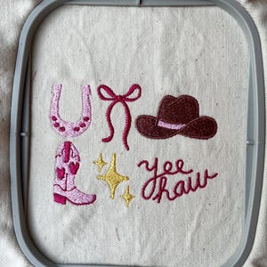 May include: Embroidered design with a pink horseshoe, a pink bow, a brown cowboy hat, a pink cowboy boot, and two yellow stars. The phrase "Yee Haw" is embroidered in pink. The design is set within a gray embroidery hoop.