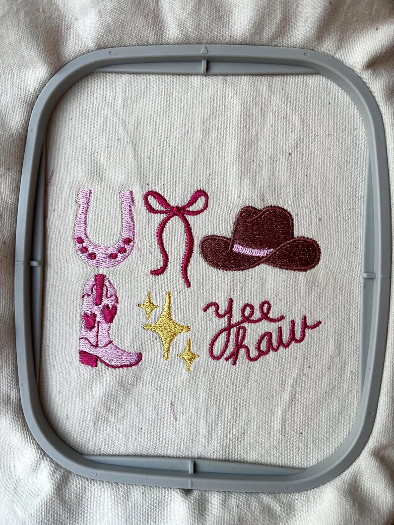 May include: Embroidered design with a pink horseshoe, a pink bow, a brown cowboy hat, a pink cowboy boot, and two yellow stars. The phrase "Yee Haw" is embroidered in pink. The design is set within a gray embroidery hoop.