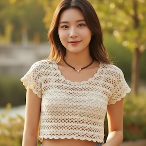 Peut inclure: Haut en crochet de couleur crème avec un décolleté festonné et des manches courtes. Le motif ajouré du haut crée un aspect délicat et aéré. Un collier marron avec un petit pendentif est également visible.