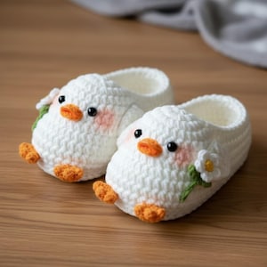 Puede incluir: Un par de zapatillas blancas de ganchillo con forma de pollitos. Las zapatillas tienen picos y patas naranjas, ojos negros y mejillas rosas. Cada zapatilla está decorada con una pequeña flor blanca y hojas verdes.