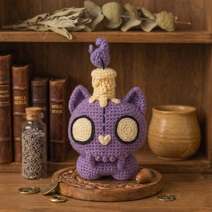 May include: A purple crocheted cat figure with large cream-colored circular eyes and a small heart-shaped nose. A cream-colored candle with a purple flame sits atop the cat's head. The figure features a stitched line down the center of its forehead.