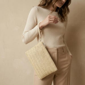 Könnte beinhalten: Rechteckige Clutch aus hellbeigem Strohgeflecht mit Reißverschluss und Handschlaufe. Die Clutch hat ein Fischgrätenmuster. Der Hintergrund ist ein neutrales Beige.