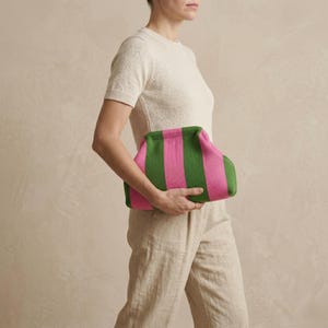 Può includere: Una pochette a righe verdi e rosa. La borsa è tenuta da una persona. La pochette ha una forma arrotondata ed è realizzata in un materiale morbido. Le strisce sono verticali e uniformemente distanziate.
