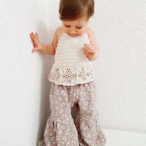 May include: A young child wearing a white crocheted halter top with a floral design and brown pants with a white leopard print.