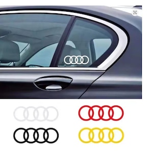 May include: A white Audi logo sticker is displayed on the rear window of a dark gray car. Below, there are four additional Audi logo stickers in white, red, black, and yellow.