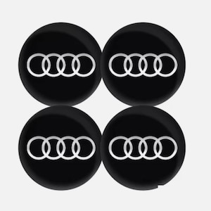 May include: Four black circular emblems with the Audi logo in white. The logo consists of four interlocking rings. The emblems are arranged in a square formation against a white background.