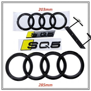 May include: A set of black automotive emblems including two four-ring logos measuring 203mm and 285mm, and two SQ5 badges with yellow accents. A black metal mounting bracket with four prongs is included to facilitate the installation of these car parts.