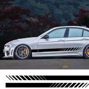 May include: Silver sedan with black racing stripes along the side. The stripes are a solid black line with a parallel design of angled lines. The car has black rims and yellow brake calipers. The background is a blurred view of trees.
