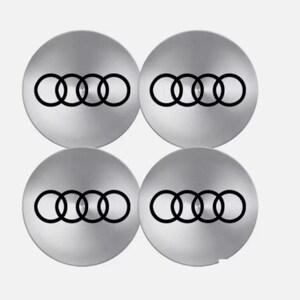 May include: Four silver-colored wheel center caps with the Audi logo, featuring four interlocking rings in black. The caps are circular and have a brushed metal appearance, set against a plain white background.