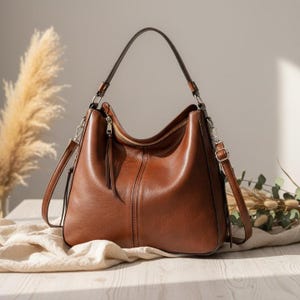 Peut inclure: Un sac à bandoulière en cuir marron avec une poignée supérieure et une bandoulière amovible. Le sac a une fermeture éclair et une couture verticale à l'avant. Le sac est posé sur un tissu blanc.