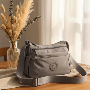 Puede incluir: Un bolso bandolera gris con múltiples compartimentos con cremallera y una correa ajustable para el hombro. El bolso presenta un emblema circular en la parte delantera y está hecho de un material liso y duradero. El bolso se exhibe sobre una mesa de madera con un jarrón de flores secas.