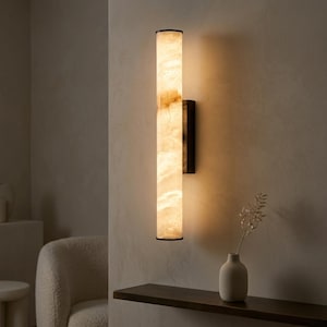 May include: A tall, rectangular wall sconce with a cream-colored, marbled appearance. The light fixture has a black frame and trim. The sconce is mounted on a neutral-colored wall. A small, white vase with a sprig of dried flowers sits on a dark shelf.