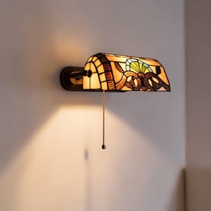 Puede incluir: Una lámpara de pared de vidrieras con acabado bronce. La lámpara presenta una pantalla rectangular con un diseño colorido de vidrio naranja, amarillo, marrón y azul. Una cadena cuelga de la parte inferior. La lámpara está montada en una pared blanca.