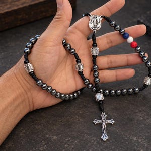 Op de afbeelding: Een rozenkrans met gepolijste donkergrijze hematietkralen aan een zwart koord, geaccentueerd met zilverkleurige metalen tussenstukjes, een centrale medaille met een religieuze figuur en een zilverkleurig kruisbeeld. Bevat drie kleine blauwe, witte en rode kralen.