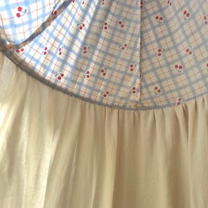 May include: A garment featuring a top section with a light blue and white checkered pattern adorned with small red cherry prints. This section is joined by a horizontal seam to a solid, off-white, gathered fabric skirt that hangs downward.